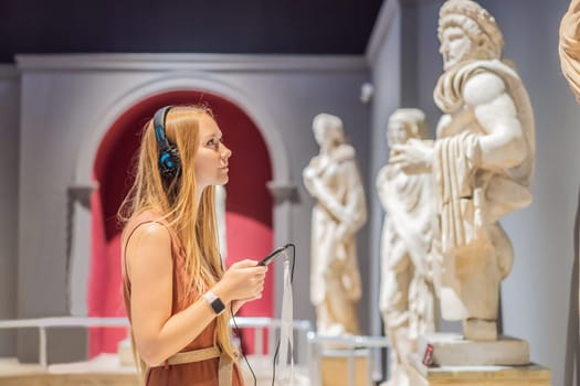 Portrait of contemporary young woman looking at sculptures and listening to audio guide at museum exhibition.