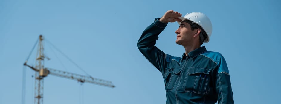 A builder in work clothes and a helmet stands on a construction site against the background of a construction crane