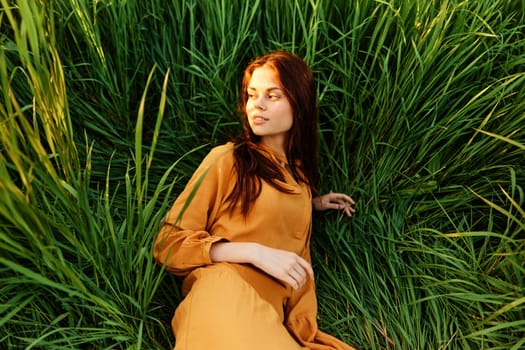 a happy woman lies in the tall green grass in a long orange dress and looks away smiling pleasantly. High quality photo