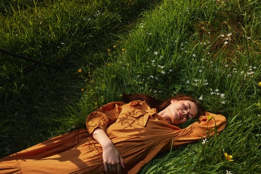 horizontal photo of a happy woman lying in the grass in a long orange dress, with her eyes closed and a pleasant smile on her face, with her hand behind her head. High quality photo