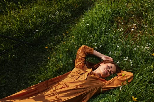 horizontal photo of a happy, relaxed woman, resting lying in the grass, in a long orange dress, with closed eyes and a pleasant smile on her face, enjoying harmony with nature and recuperating. High quality photo