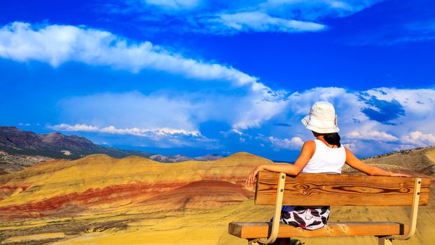 Young woman sitting on the hill-top bench relaxing while watching the colorful Painted hills at John Day Fossil Beds National Monument, Oregon