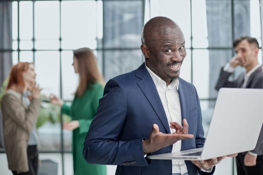 Young African American man in a business suit holding a laptop against the background of his colleagues