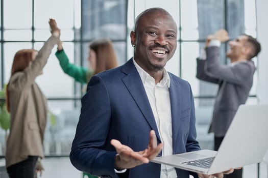Young African American man in a business suit holding a laptop against the background of his colleagues