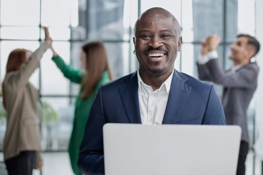 Young African American man in a business suit holding a laptop against the background of his colleagues