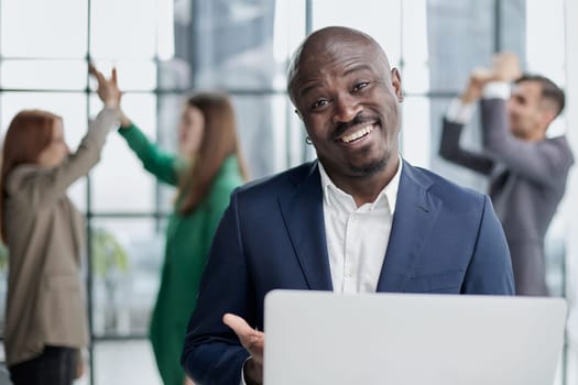 Young African American man in a business suit holding a laptop against the background of his colleagues