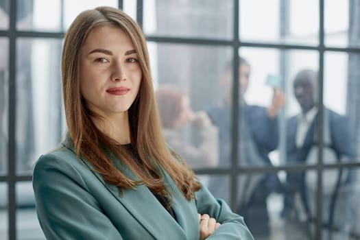 businesswoman standing with her arms crossed in an office while her colleagues have a meeting in the background.