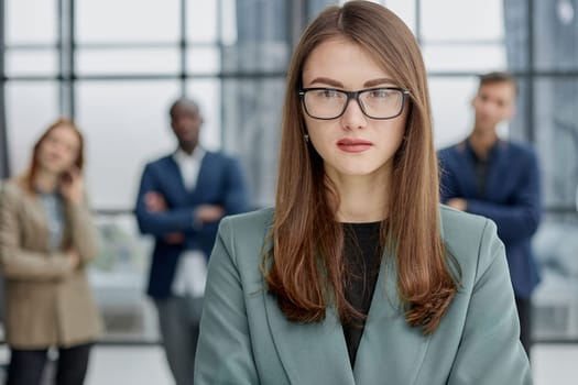 businesswoman standing with her arms crossed in an office while her colleagues have a meeting in the background.