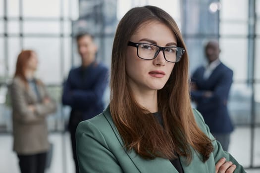 businesswoman standing with her arms crossed in an office while her colleagues have a meeting in the background.