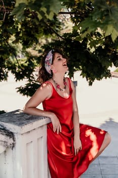 woman in a red silk dress and a bandage on her head smiles against the background of the leaves of a tree. She is leaning on the coop and looking into the camera. Vertical photo