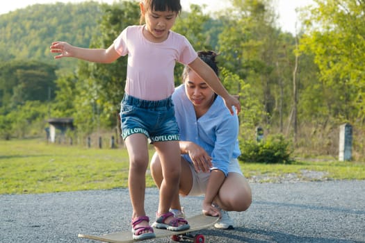 Mother teaching her daughter how to skateboard in the park. Child riding skate board. Healthy sports and outdoor activities for school children in the summer.