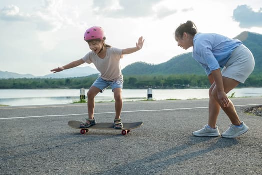 Mother teaching her daughter how to skateboard in the park. Child riding skate board. Healthy sports and outdoor activities for school children in the summer.