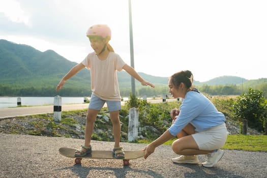 Mother teaching her daughter how to skateboard in the park. Child riding skate board. Healthy sports and outdoor activities for school children in the summer.