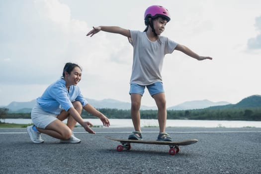 Mother teaching her daughter how to skateboard in the park. Child riding skate board. Healthy sports and outdoor activities for school children in the summer.