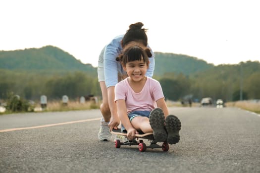 Mother teaching her daughter how to skateboard in the park. Child riding skate board. Healthy sports and outdoor activities for school children in the summer.
