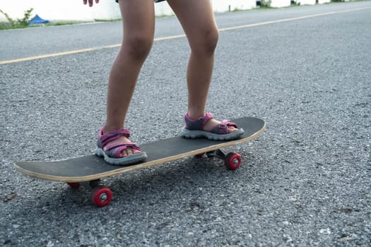 Cute little girl learns how to skateboard at outdoor street, focus on legs standing on board. Healthy sports and outdoor activities for school children in the summer.