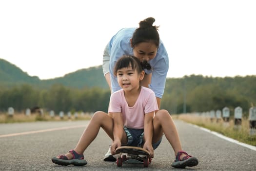 Mother teaching her daughter how to skateboard in the park. Child riding skate board. Healthy sports and outdoor activities for school children in the summer.