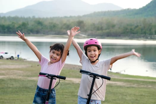 Two cute little girls smiling and posing together in summer garden. Happy kid riding kick scooter in the park. Healthy sports and outdoor activities for children.