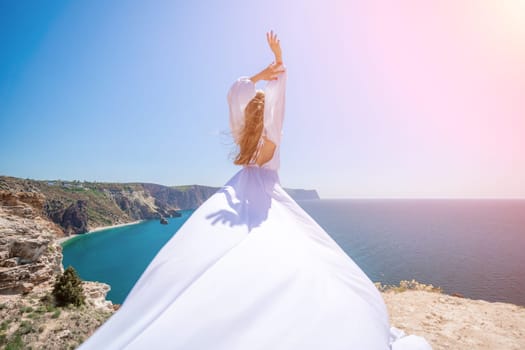 woman sea white dress. Blonde with long hair on a sunny seashore in a white flowing dress, rear view, silk fabric waving in the wind. Against the backdrop of the blue sky and mountains on the seashore