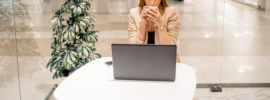 A business woman sits in a cafe, works at a computer, drinks coffee. She is wearing a beige jacket, brown top and black trousers