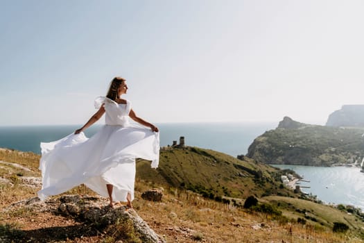 Happy woman in a white dress and hat stands on a rocky cliff above the sea, with the beautiful silhouette of hills in thick fog in the background