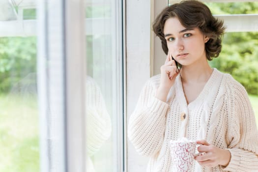 Portrait lonely caucasian young woman sitting near window with phone in her hand in a white knitted sweater, spending time in the morning on the veranda in a country house