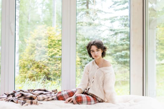 Portrait lonely caucasian young woman sitting near window apartments in a white sweater and a red plaid, spends time in the morning on the veranda in a country house