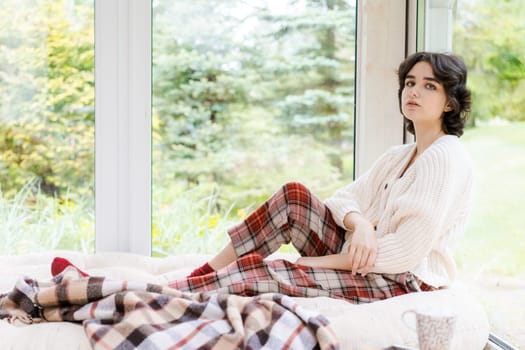 Portrait lonely caucasian young woman sitting near window apartments in a white sweater and a red plaid, spends time in the morning on the veranda in a country house