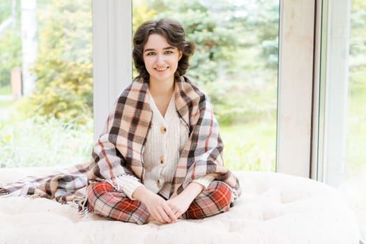 Portrait lonely caucasian young woman sitting near window apartments in a white sweater and a red plaid, spends time in the morning on the veranda in a country house