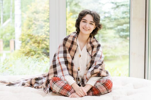 Portrait lonely caucasian young woman sitting near window apartments in a white sweater and a red plaid, spends time in the morning on the veranda in a country house