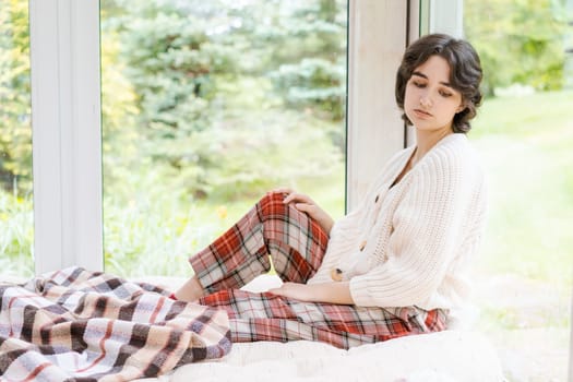 Portrait lonely caucasian young woman sitting near window apartments in a white sweater and a red plaid, spends time in the morning on the veranda in a country house