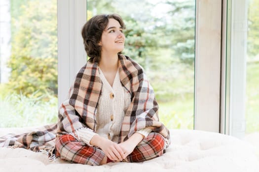 Portrait lonely caucasian young woman sitting near window apartments in a white sweater and a red plaid, spends time in the morning on the veranda in a country house