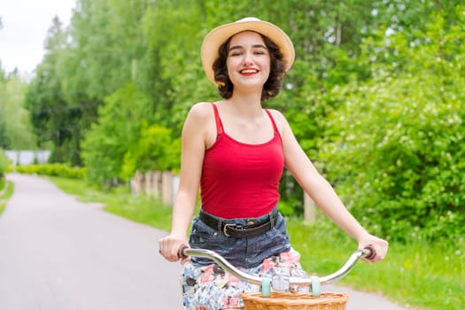 Portrait beautiful young girl in hat with bicycle on street in countryside in sunlight outdoor in a red tank top on a summer day rides on the weekend