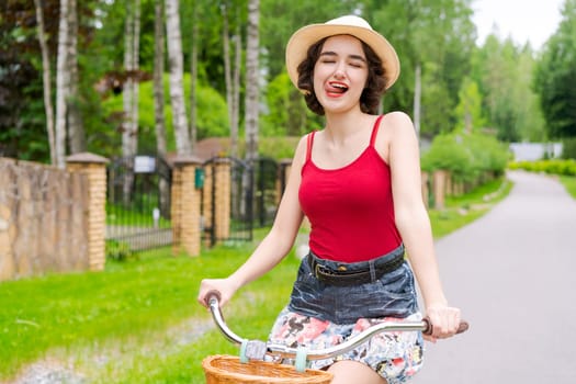 Portrait beautiful young girl in hat with bicycle on street in countryside in sunlight outdoor in a red tank top on a summer day rides on the weekend
