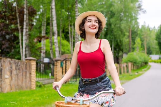 Portrait beautiful young girl in hat with bicycle on street in countryside in sunlight outdoor in a red tank top on a summer day rides on the weekend