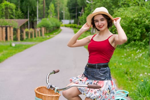 Portrait beautiful young girl in hat with bicycle on street in countryside in sunlight outdoor in a red tank top on a summer day rides on the weekend