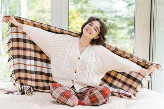 Portrait lonely caucasian young woman sitting near window apartments in a white sweater and a red plaid, spends time in the morning on the veranda in a country house