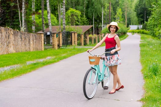 Portrait beautiful young girl in hat with bicycle on street in countryside in sunlight outdoor in a red tank top on a summer day rides on the weekend