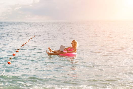 Woman works on laptop in sea. Freelancer, blond woman in sunglases floating on an inflatable big pink donut with a laptop in the sea at sunset. People summer vacation rest lifestyle concept