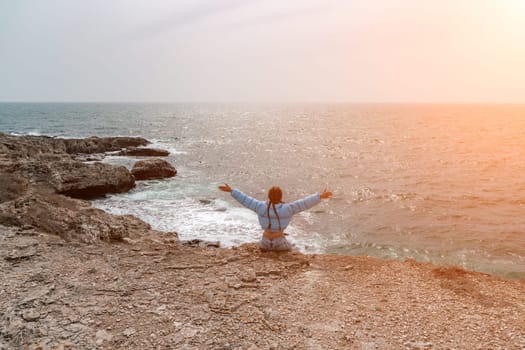 woman sea travel. A woman in a blue jacket sits on a rock above a cliff above the sea, looking at the stormy ocean. Girl traveler rests, thinks, dreams, enjoys nature.