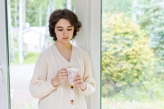 Female portrait on background window. Young woman sitting on the veranda drinking her hot coffee. Cozy winter morning. Peace of mind and mental health. In a bright knitted sweater