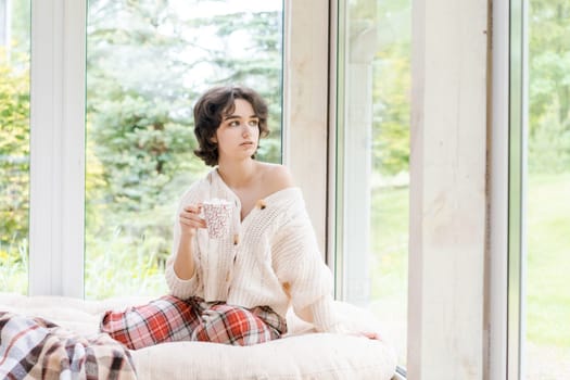 Female portrait on background window. Young woman sitting on veranda drinking her hot coffee. Cozy winter morning. Peace of mind and mental health. In a bright knitted sweater