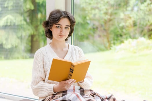 Brunette woman in warm sweater reading book sitting on room on the veranda by the window looking mood