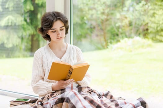 Brunette woman in warm sweater reading book sitting on room on the veranda by the window looking mood