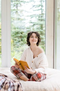 Brunette woman in warm sweater reading book sitting on room on the veranda by the window looking mood