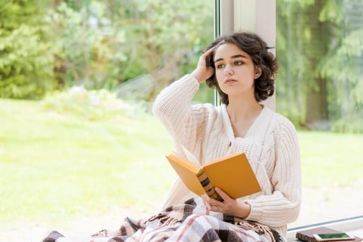 Brunette woman in warm sweater reading book sitting on room on the veranda by the window looking mood