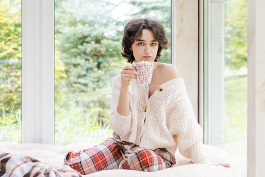 Female portrait on background window. Young woman sitting on veranda drinking her hot coffee. Cozy winter morning. Peace of mind and mental health. In a bright knitted sweater