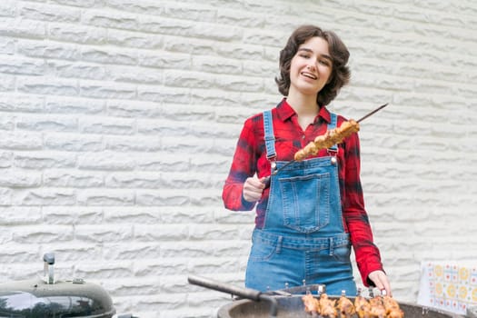 Barbeque party in garden with young woman in denim overalls and red plaid shirt in a country house on the terrace preparing meat skewers