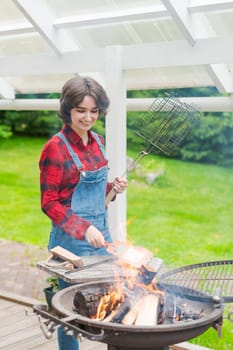 Barbeque party in garden with young woman in denim overalls and red plaid shirt in a country house on the terrace preparing meat skewers