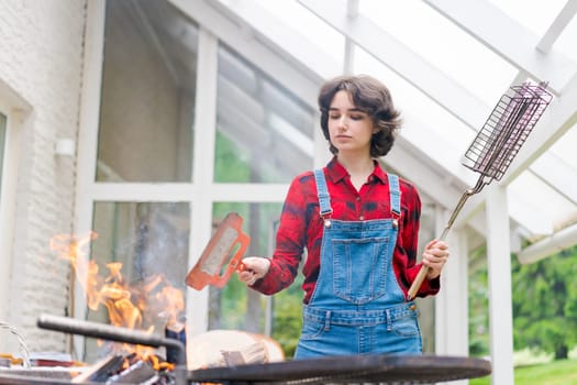 Barbeque party in garden with young woman in denim overalls and red plaid shirt in a country house on the terrace preparing meat skewers